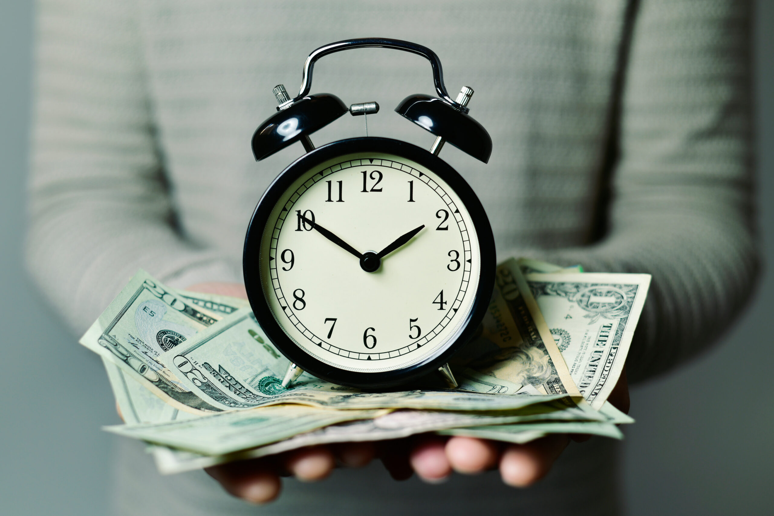 A person holding an alarm clock on a pile of money—time is money!